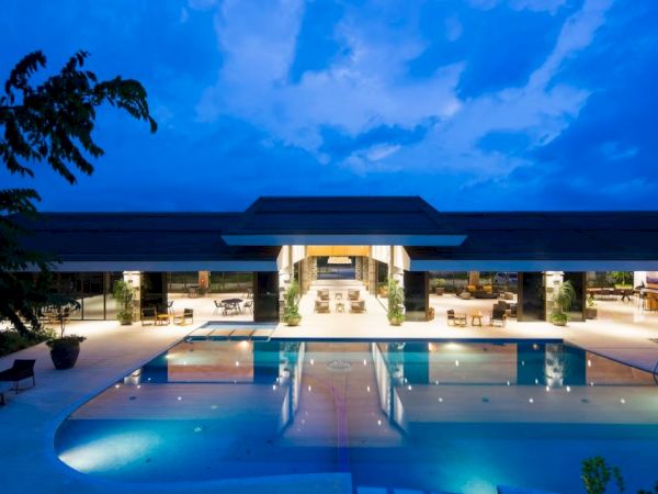 A luxurious, modern building with large windows is shown at dusk, featuring a well-lit swimming pool surrounded by lounge chairs.