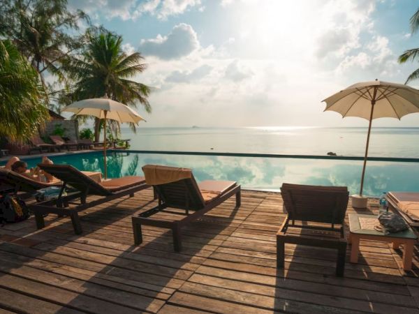 A serene poolside with lounge chairs, umbrellas, palm trees, and a beautiful ocean view under a partly cloudy sky.
