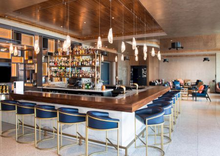 A modern bar with blue chairs, wooden counter, hanging lights, and a stocked liquor shelf. Lounge seating is visible in the background.