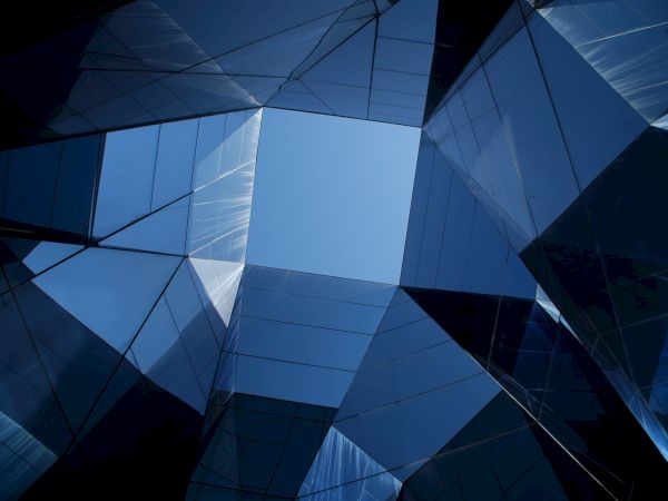 The image shows an upward view of geometric, mirrored buildings surrounding a clear blue sky, creating a modern architectural pattern.