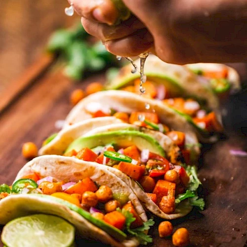 A row of vegetable tacos on a wooden board with a hand squeezing lime juice over them, garnished with cilantro and lime slices.