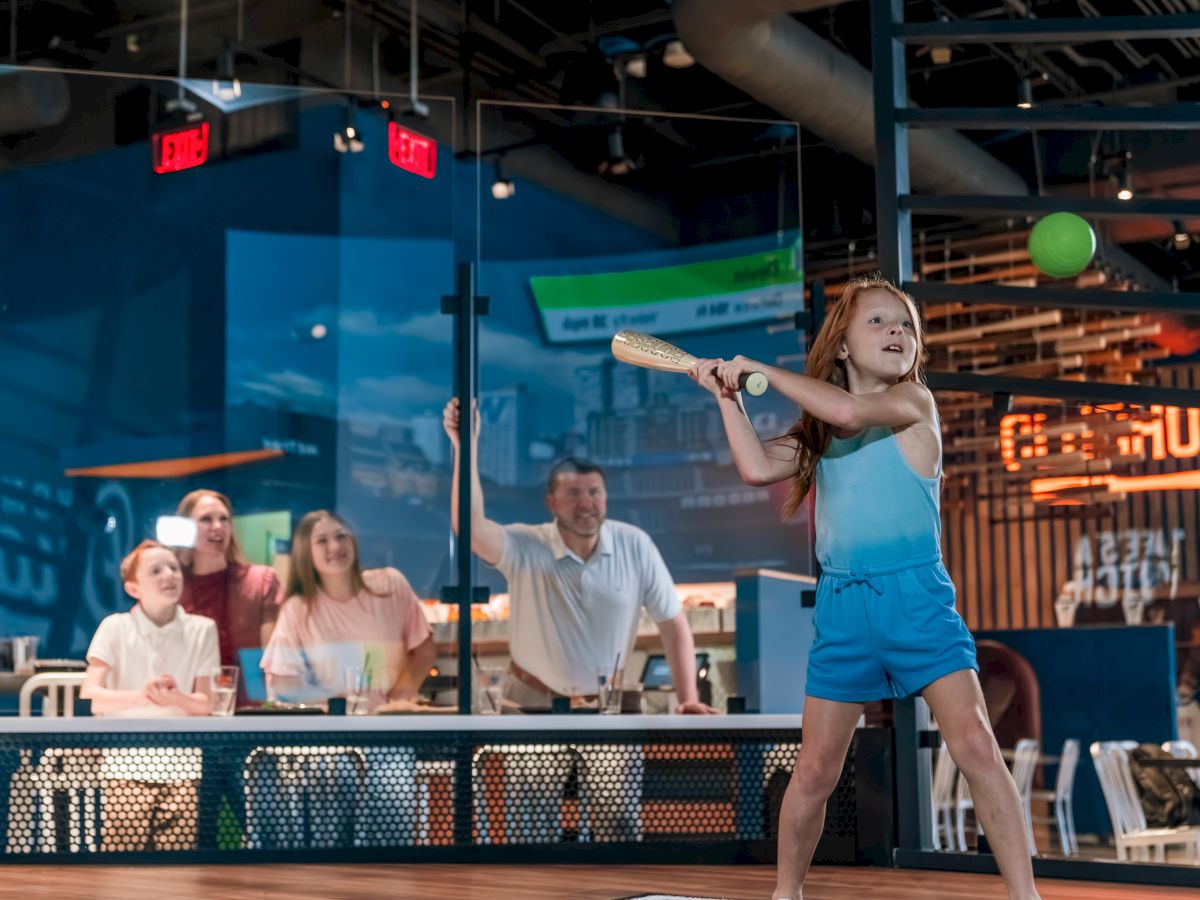 A child is playing baseball indoors, preparing to hit the ball with a bat, while a group of people watches in the background.