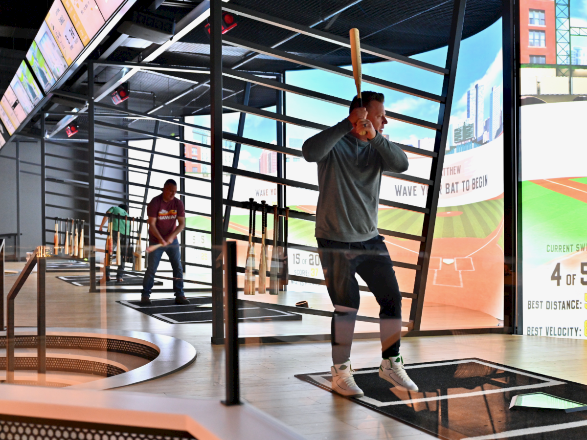 People are at an indoor batting cage with digital displays showing scores and stats in real-time, enhancing the baseball experience.