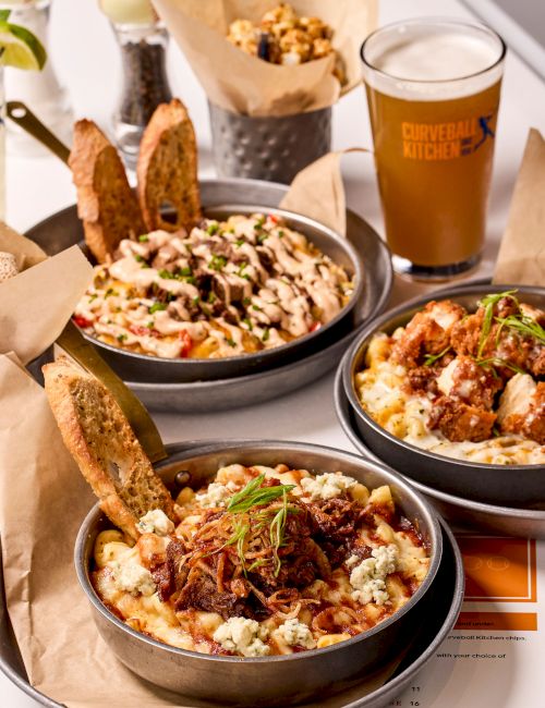 The image shows bowls of savory dishes with meat and bread, a glass of beer, popcorn in a metal cup, and baseball-themed decor.