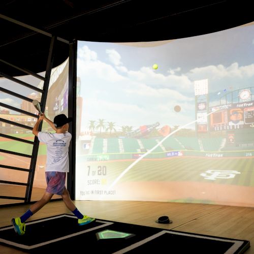 A person is playing a virtual baseball game, swinging a bat at a screen displaying a baseball field simulation.