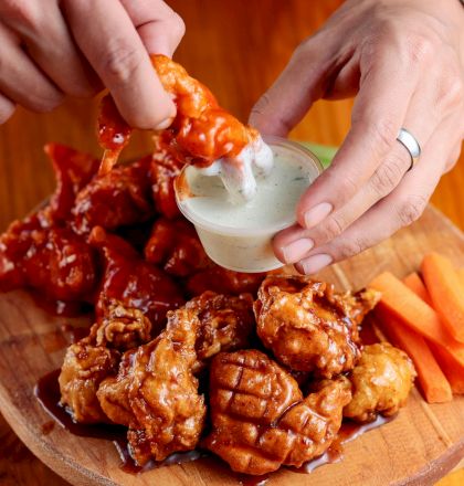 A person dips a saucy chicken piece into a creamy dressing, on a wooden platter with more chicken and carrot sticks.