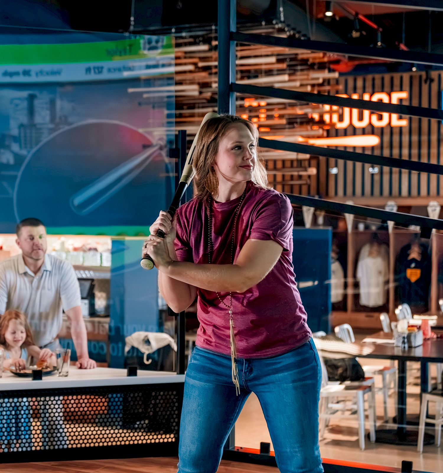 A person holding a baseball bat is preparing to swing while a group watches from the background at an indoor venue.