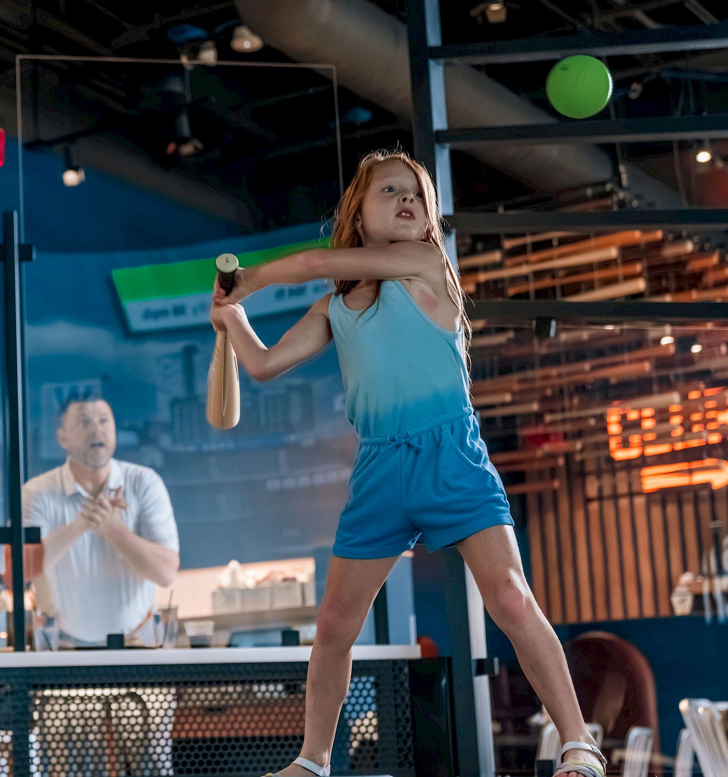 A girl swinging a bat indoors while three people cheer her on from a seating area.