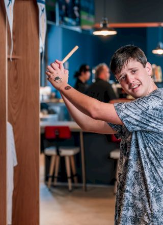 A person holding a pen in a playful pose near a wooden booth in a lively, casual indoor setting with others around.
