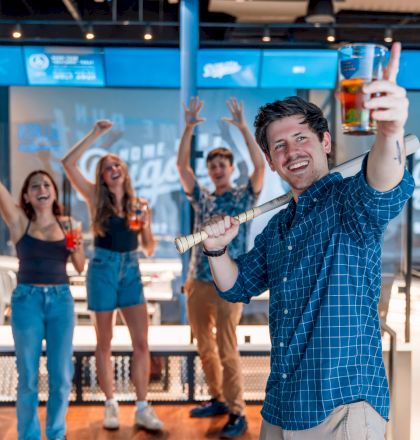 A group of friends in casual attire are enjoying a day indoors, holding drinks and celebrating, with one person in front posing with a golf club.