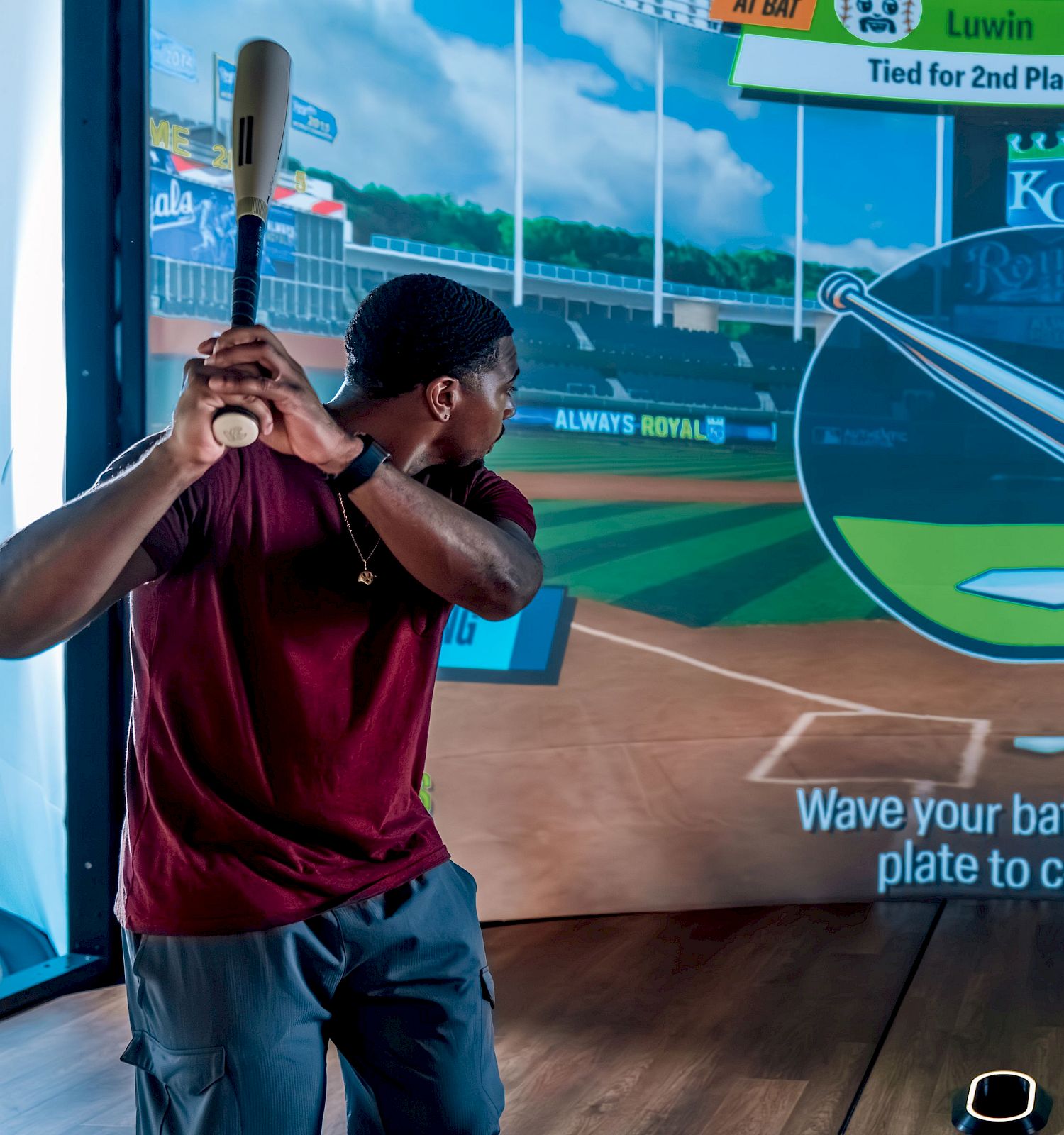 A person is playing a baseball simulation game, holding a bat and standing in front of a large screen displaying a virtual field and instructions.