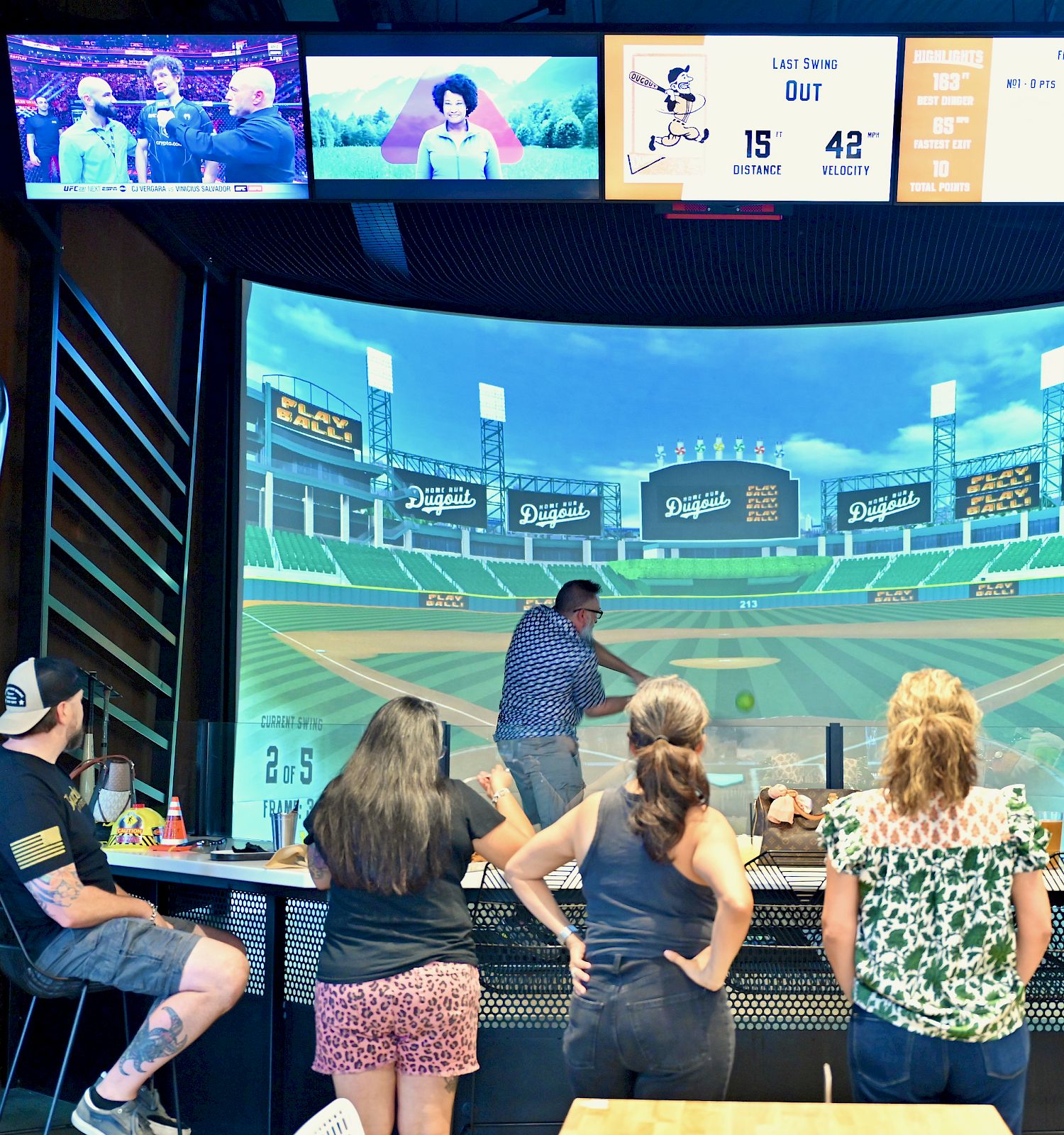 People are playing a baseball simulator game indoors, with a virtual field displayed on a large screen and scores shown above.