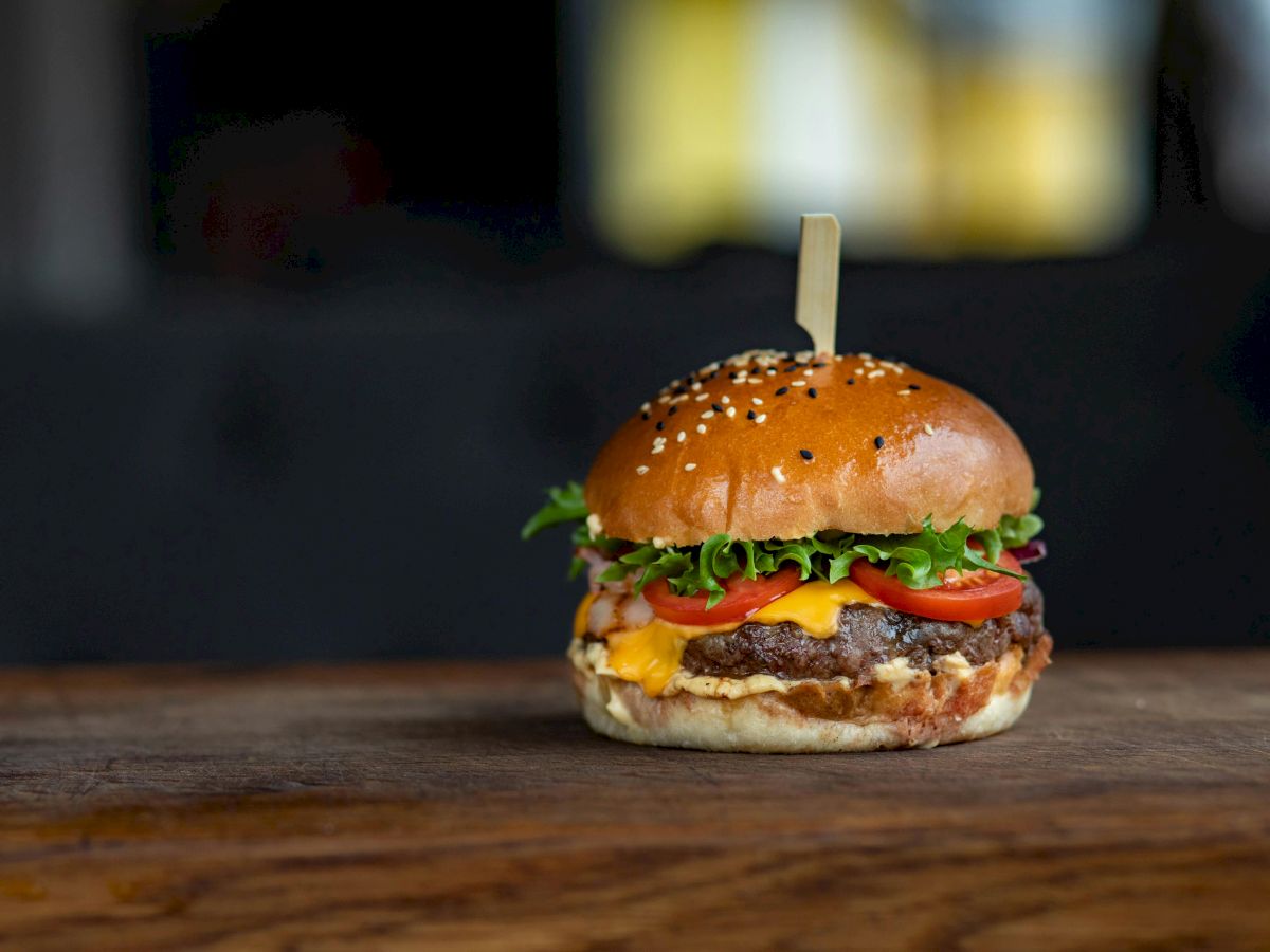 A hamburger with a sesame seed bun, cheese, meat patty, lettuce, and tomato, secured with a skewer, on a wooden surface.