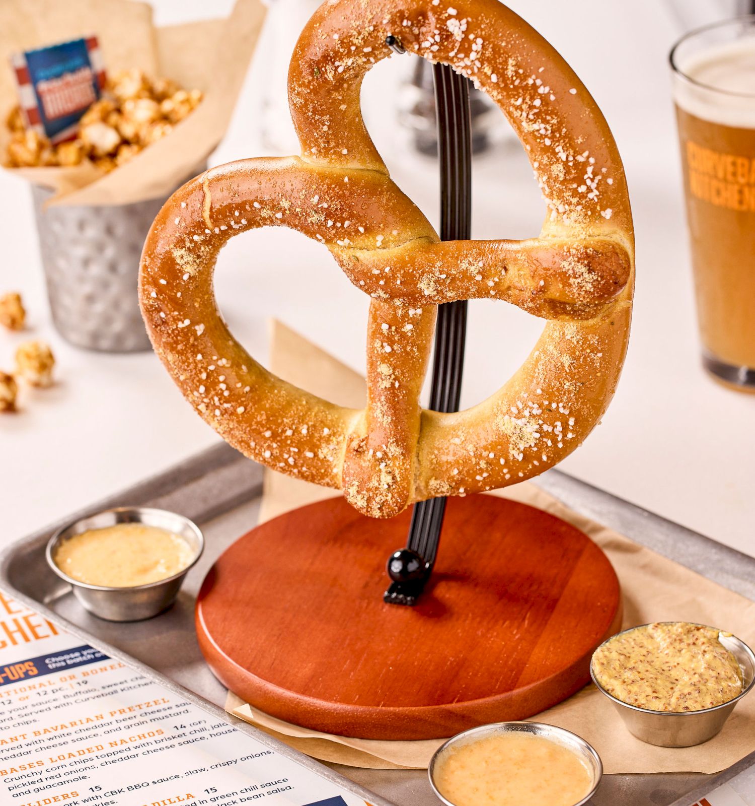 A large pretzel on a stand with three dips, popcorn, a drink, and a menu are on a table.
