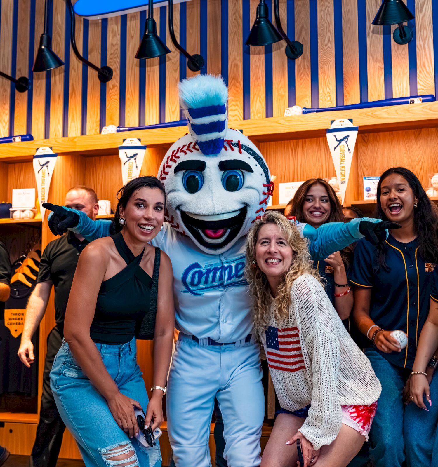 People pose with a blue mascot inside a lively venue, smiling and hugging, surrounded by fans in casual outfits and blue accents.