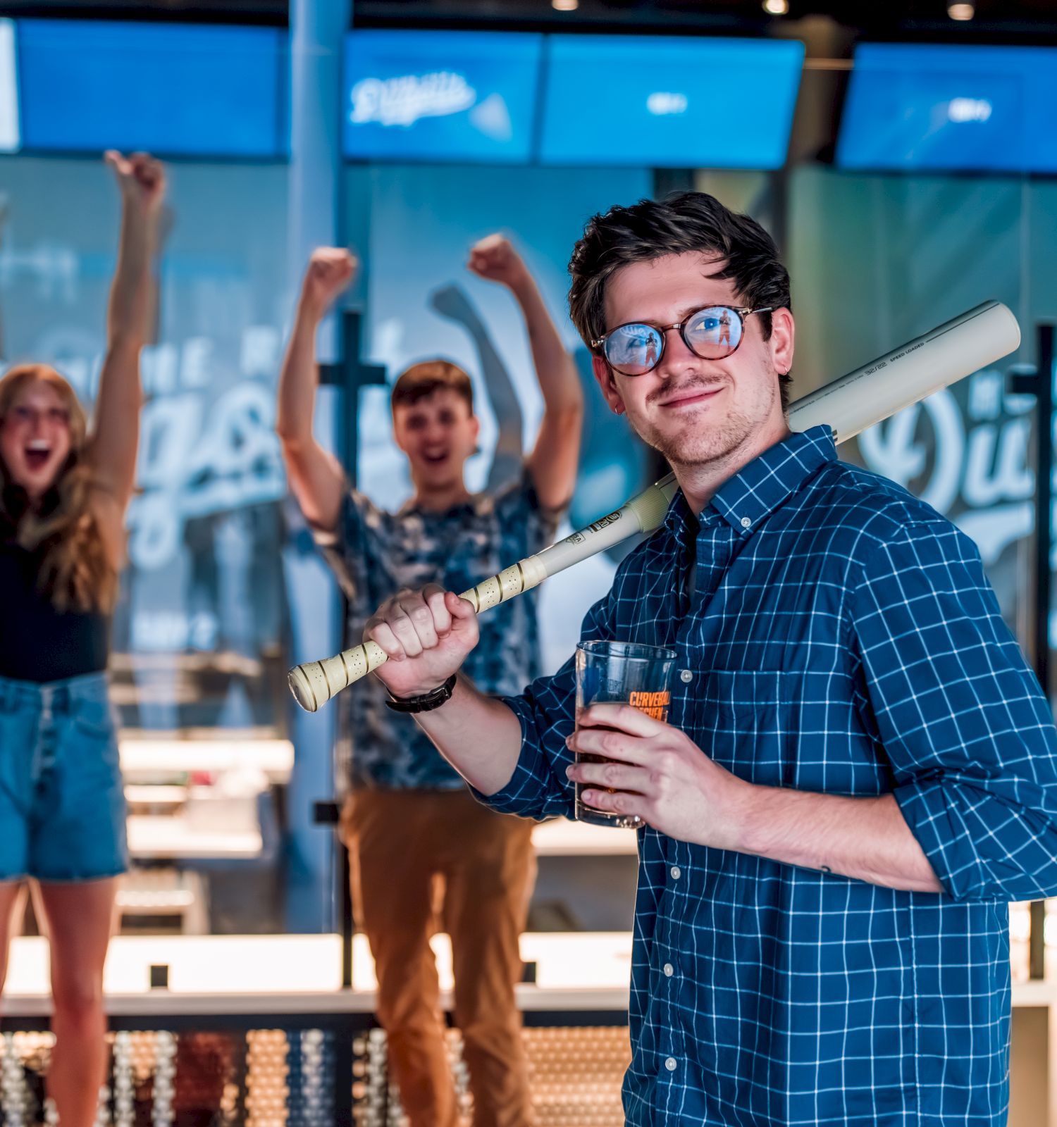 Three people are in a bar; one holds a bat and drink while two others cheer with drinks raised high.