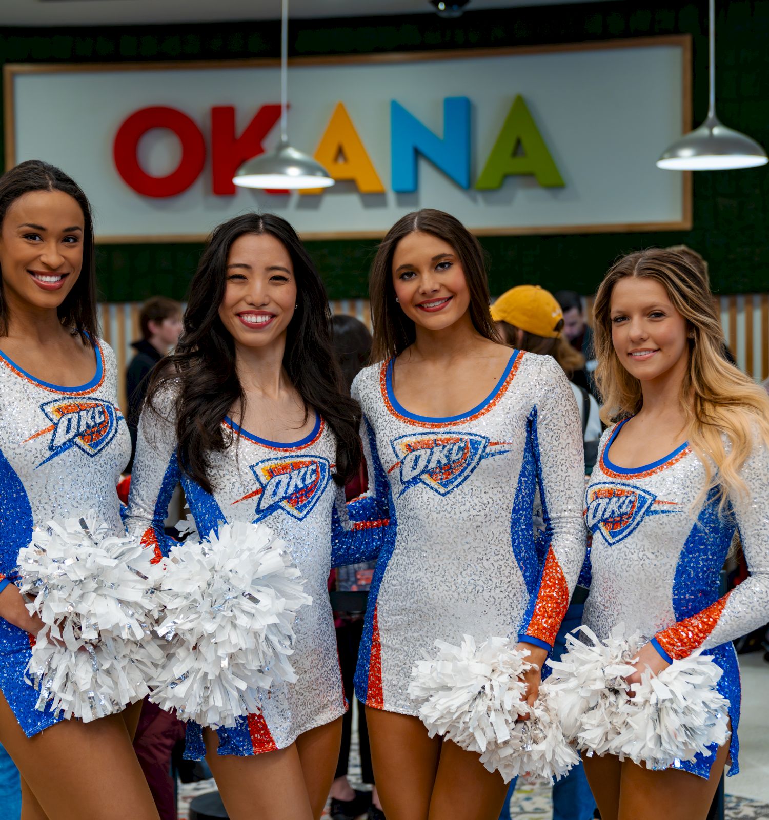 Four cheerleaders in Oklahoma City Thunder outfits posing with pom-poms in front of an &ldquo;OKCNA&rdquo; sign, smiling at the camera.