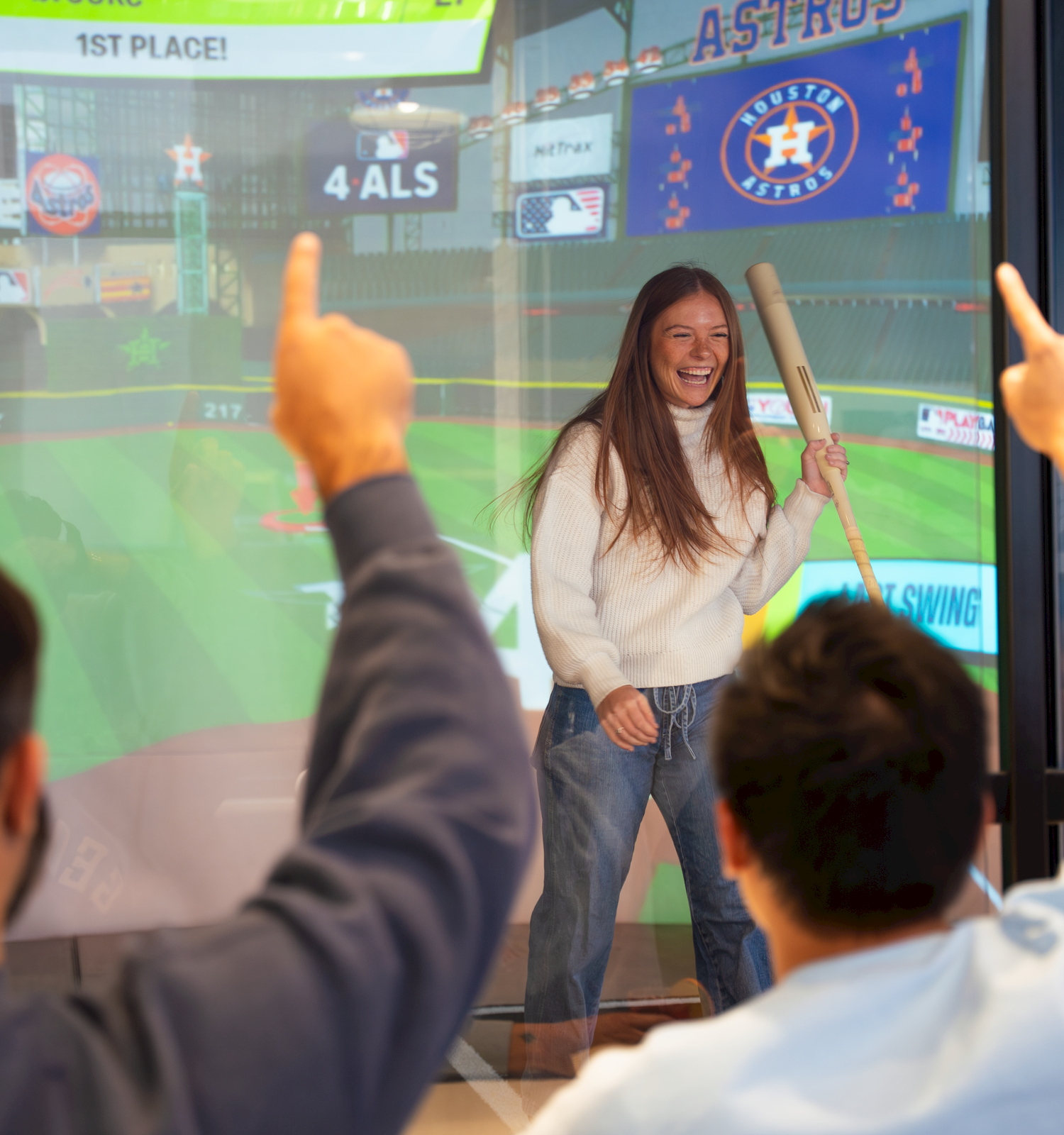 A young woman stands on-stage holding a cricket bat, while two people in the foreground gesture; scoreboard and Astros banner visible in the background.