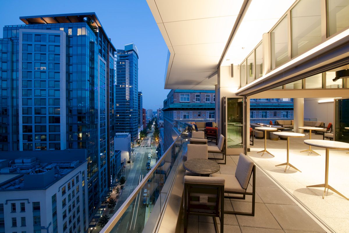 A modern urban balcony with seating overlooks city buildings and streets at dusk, with a sleek interior visible through open doors.