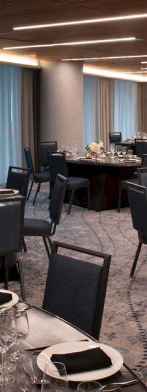 The image shows a banquet hall with round tables, set with plates, glasses, napkins, and floral centerpieces, under modern lighting.