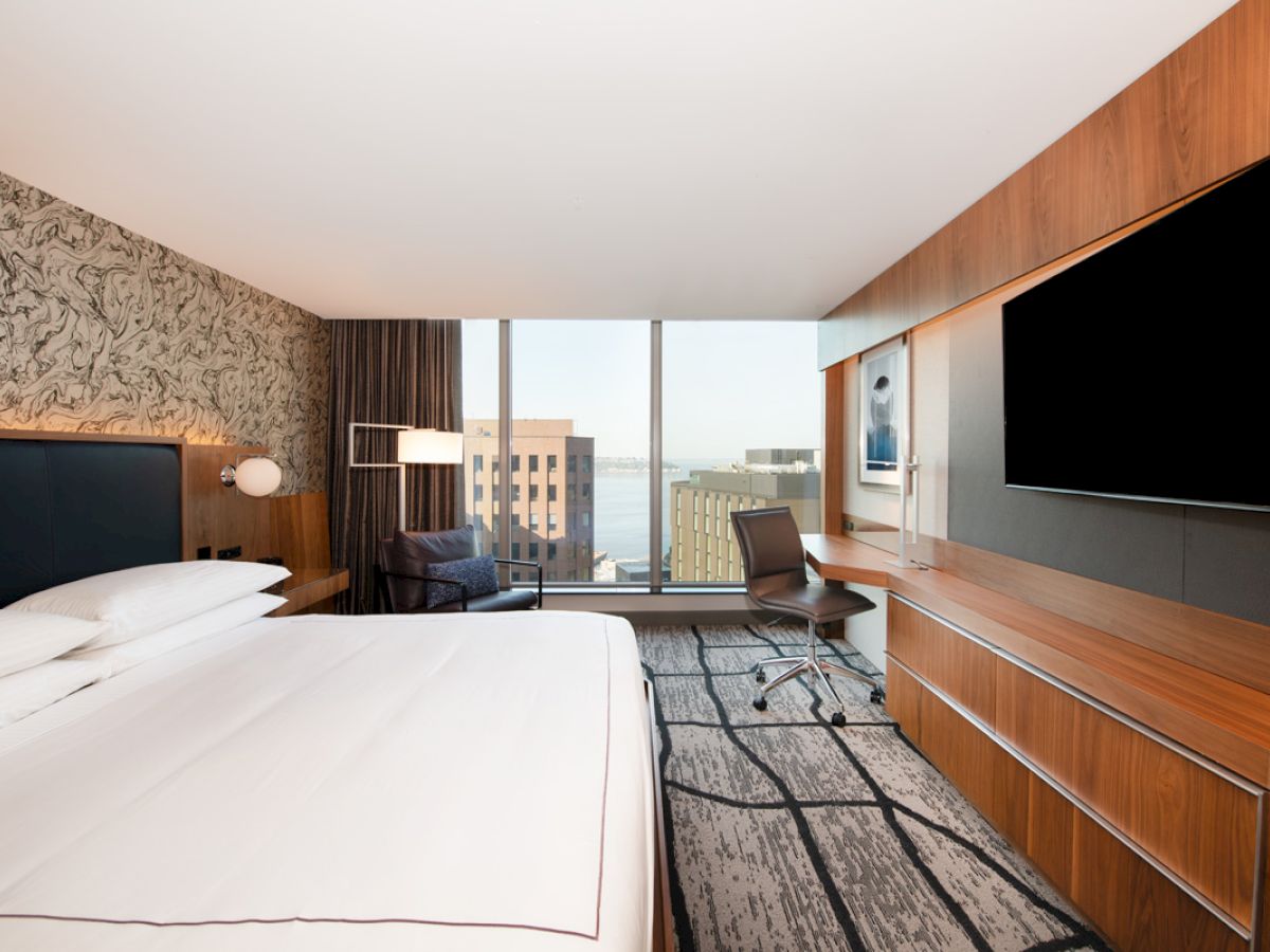 A modern hotel room with a large bed, desk, and TV, featuring a cityscape view through a window.