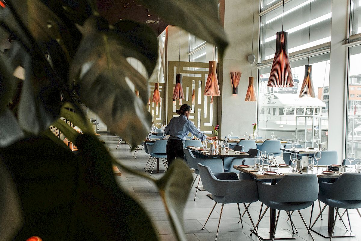 A modern restaurant interior with a worker setting tables, accented by stylish lighting and large plant leaves in the foreground.