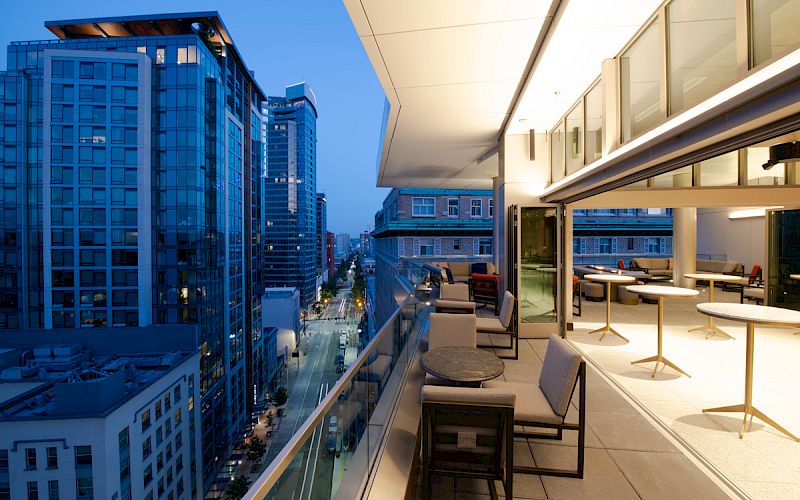 The image shows a modern rooftop terrace with seating and tables, overlooking a city street lined with tall buildings at dusk.